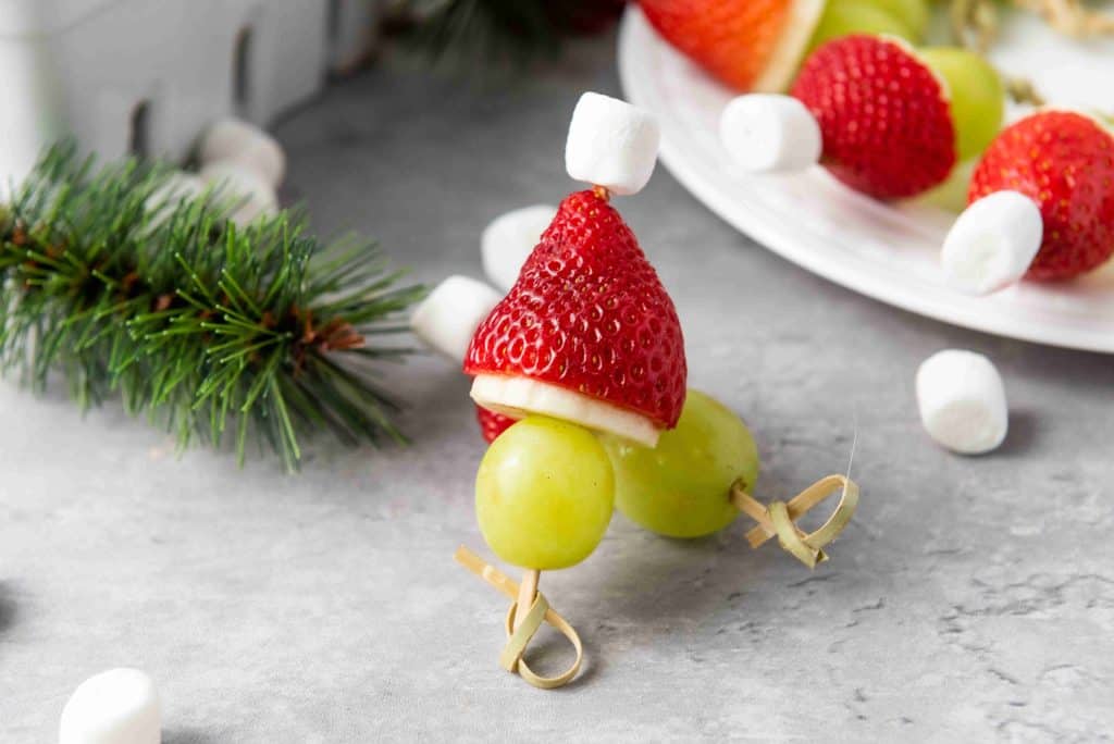A festive snack, these Grinch Fruit Kabobs feature green grapes, a banana slice, and a strawberry topped with a mini marshmallow to resemble a Christmas gnome, arranged on a gray surface with pine sprigs and marshmallows nearby.