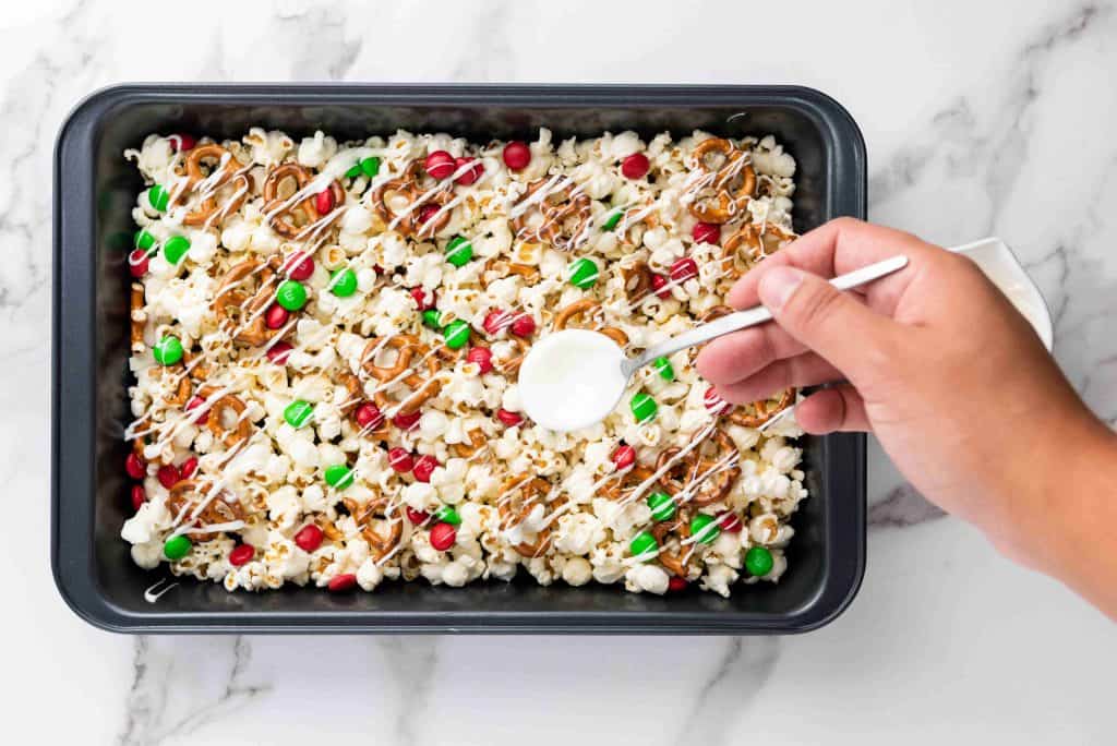 A hand drizzles white icing over a tray of holiday popcorn mix with pretzels, red and green candies, and popcorn in a black baking dish on a marble surface, creating the perfect festive treat.