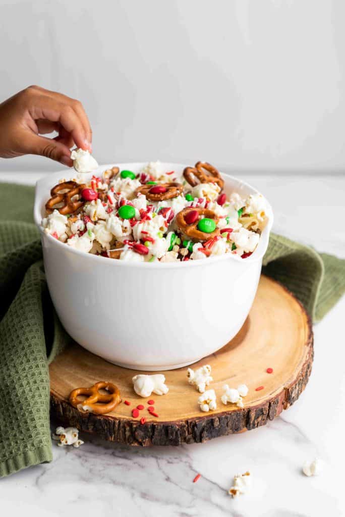 A hand reaches into a white bowl filled with holiday popcorn snack mix—popcorn, pretzels, red and green candies, and sprinkles—set on a wooden slab with a green towel atop a marble surface.