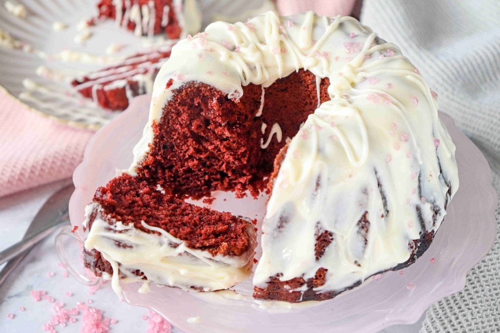 A Christmas Red Velvet Bundt Cake with white icing drizzled on top sits on a pink cake stand. One large slice is pulled out, showing the moist red interior, while pink sprinkles decorate both the festive cake and plate.