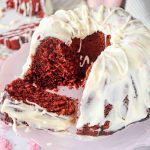 A Christmas Red Velvet Bundt Cake with white icing drizzled on top sits on a pink cake stand. One large slice is pulled out, showing the moist red interior, while pink sprinkles decorate both the festive cake and plate.
