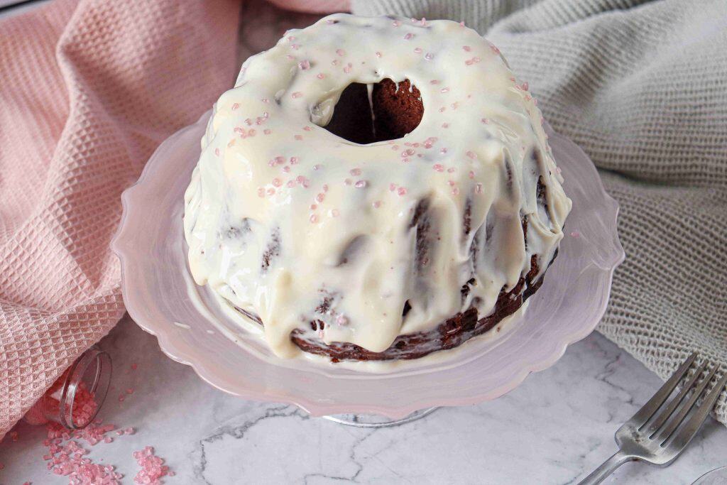 A Christmas Red Velvet Bundt Cake with white icing and pink sprinkles sits on a pink glass cake stand, surrounded by textured pink and gray cloths on a marble surface. A fork lies nearby.