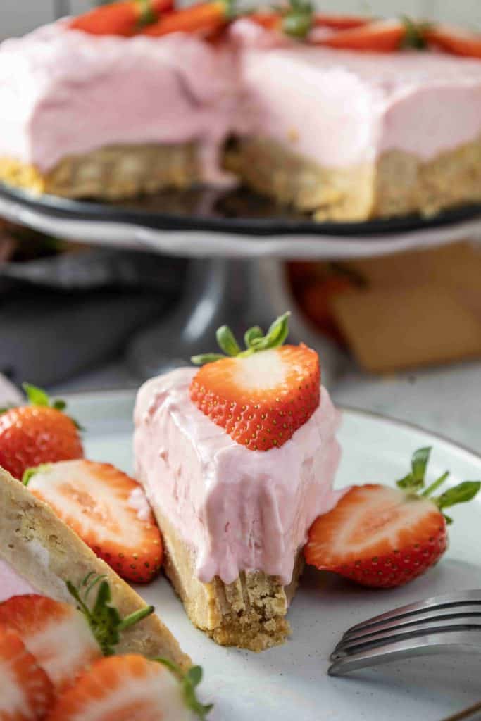 A slice of Christmas strawberry cheesecake with a pink creamy topping and fresh strawberry slices on top and on the side, served on a white plate with a fork. The rest of the festive cheesecake is visible in the background.
