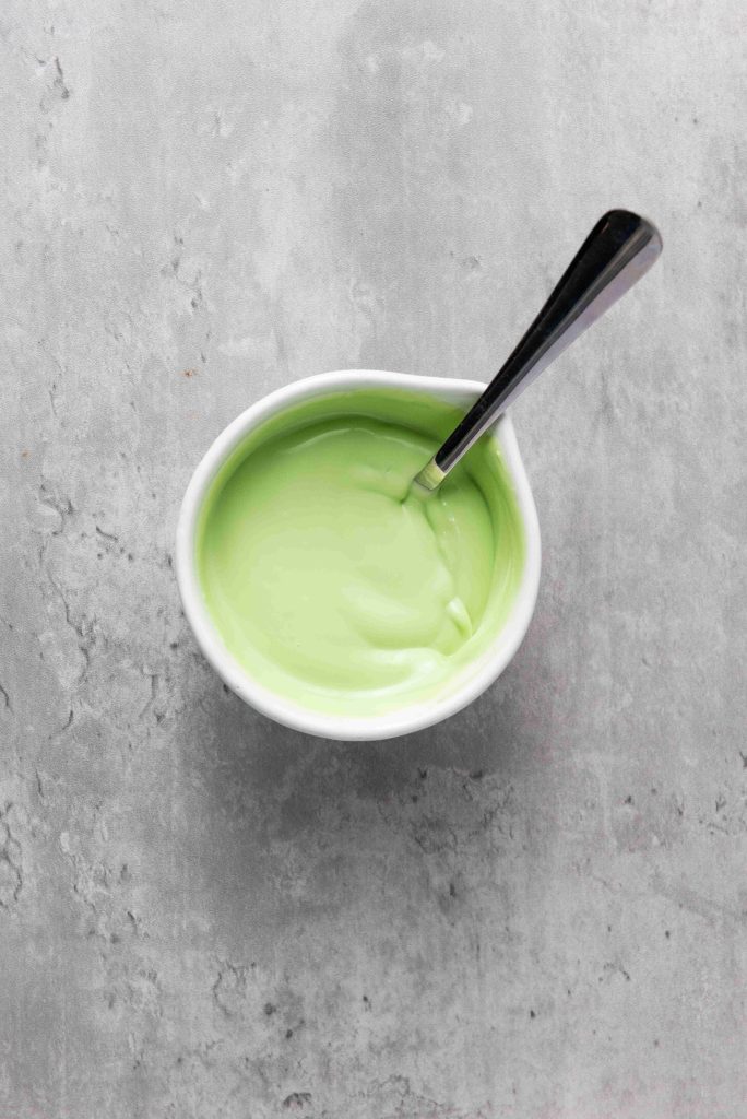A small white bowl filled with smooth, light green cream for Grinch Heart Pretzels sits on a gray textured surface. A metal spoon rests inside the bowl.