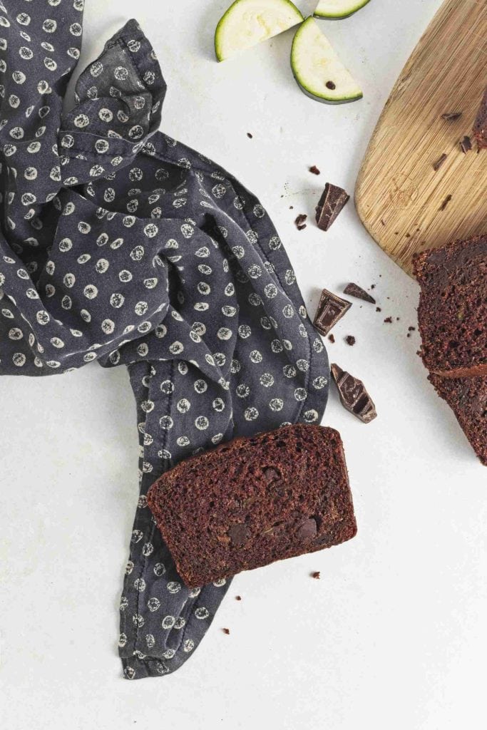 A slice of Christmas zucchini bread rests on a black and white polka dot cloth, with more bread on a wooden board, chocolate chunks, and zucchini slices nearby on a white surface.
