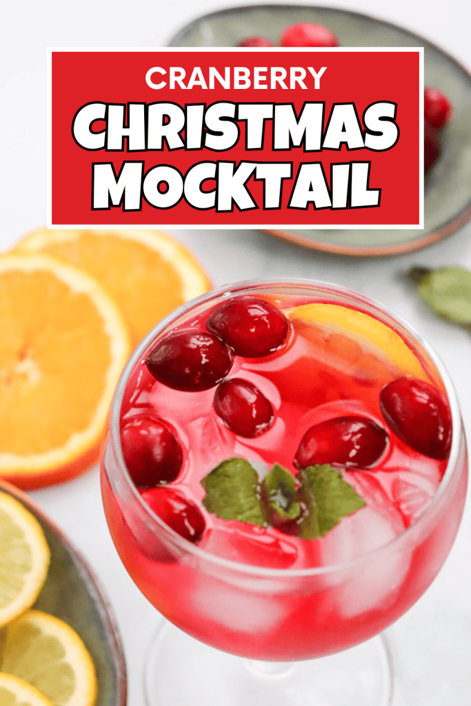 A glass of bright pink cranberry Christmas mocktail topped with ice, cranberries, orange slices, and mint leaves, with orange slices and cranberries in bowls nearby. Bold text above reads “Cranberry Christmas Mocktail.”.