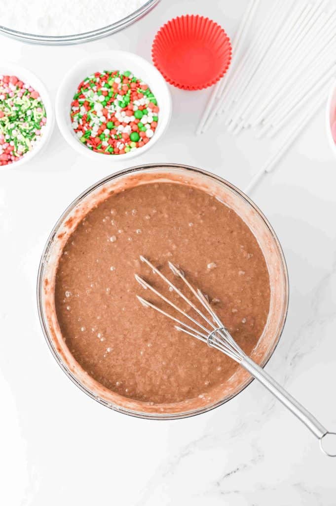 A mixing bowl filled with chocolate cake batter and a metal whisk. Nearby are bowls of colorful Christmas sprinkles, red cupcake liners, and white cake pop sticks—everything you need to make festive christmas tree cupcakes on a marble countertop.