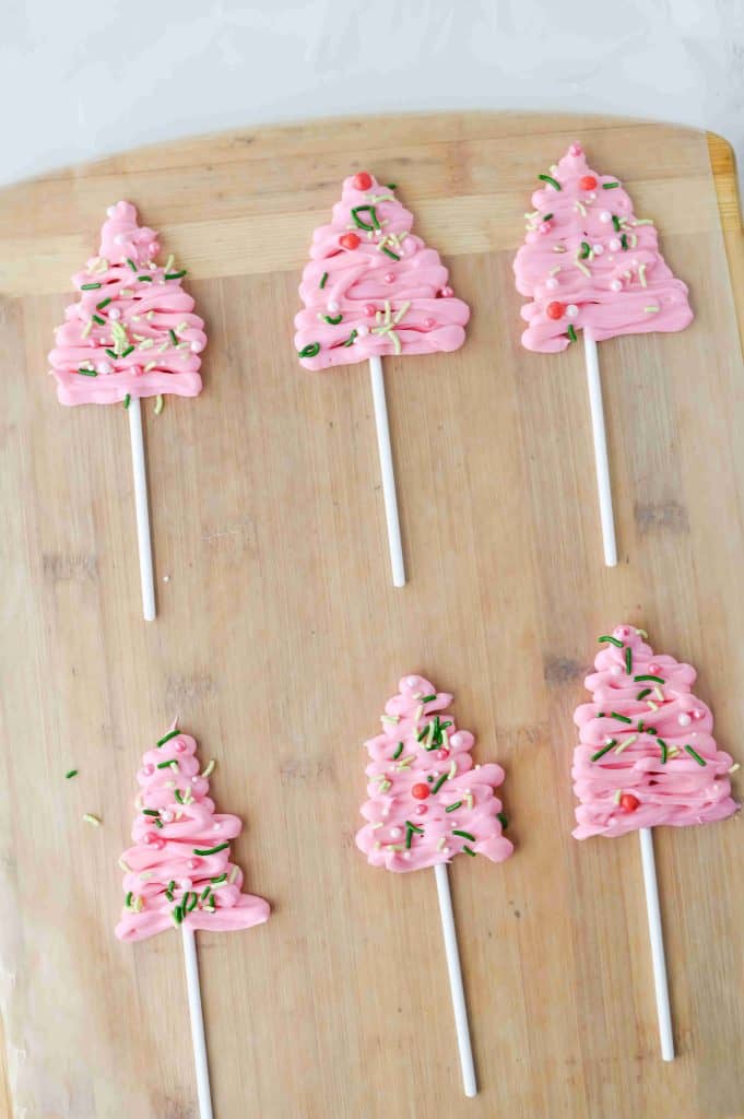Six pink Christmas tree cupcakes on white sticks are arranged on a wooden surface. Each treat is decorated with sprinkles and resembles a festive holiday lollipop or candy pop.