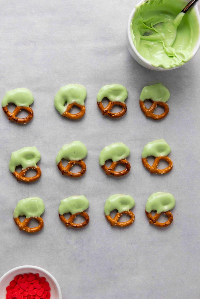 A dozen Grinch Heart Pretzels, partially dipped-pretzels-for-christmas in green coating, are lined up on parchment paper. Nearby, a bowl of green coating with a spoon and a dish of red heart-shaped sprinkles complete the festive setup.