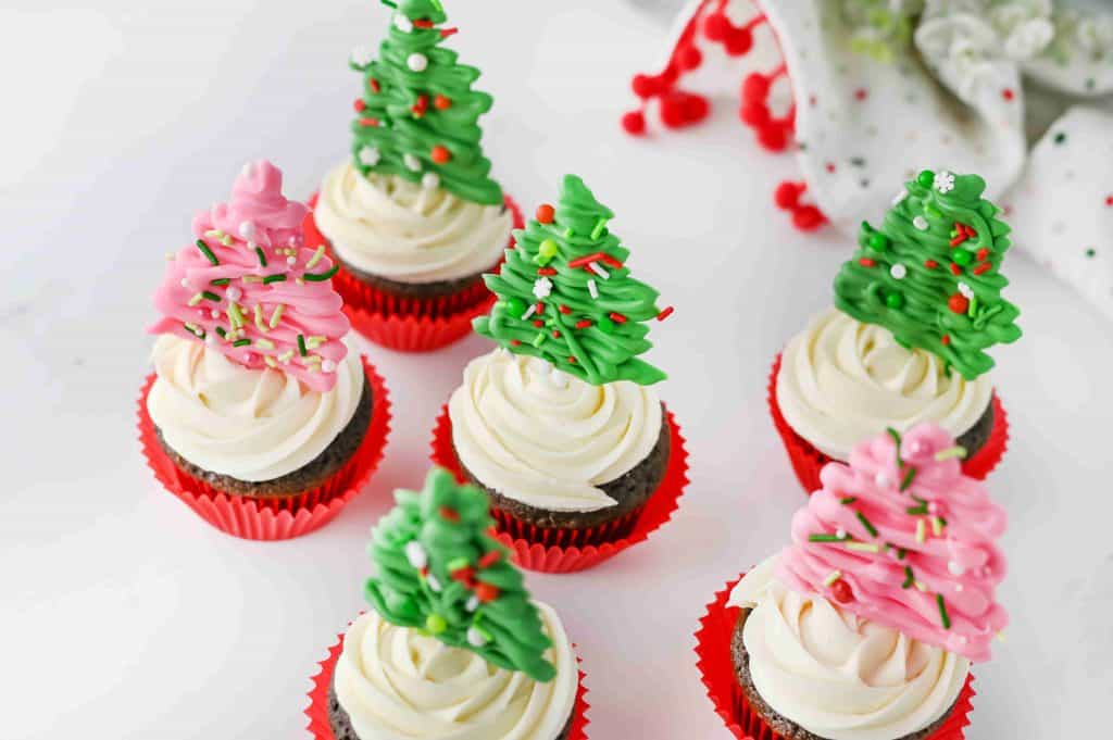 Seven christmas tree cupcakes with white frosting in red wrappers are topped with green and pink candy Christmas trees decorated with sprinkles, set on a white surface with festive decorations in the background.