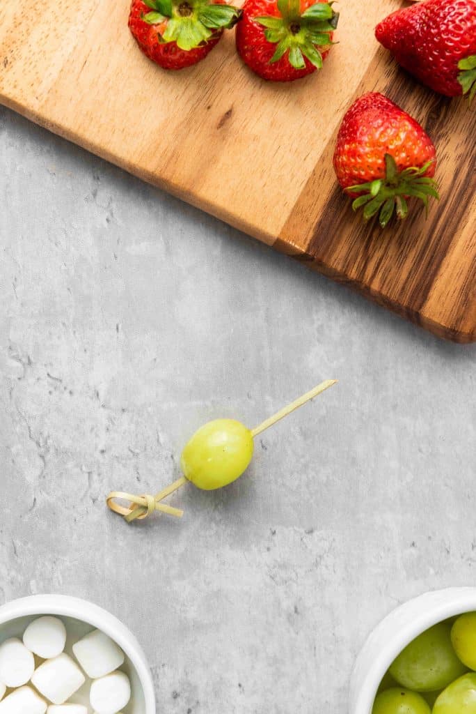 A single green grape on a toothpick sits on a gray surface, surrounded by fresh strawberries and bowls of mini marshmallows—perfect ingredients for making festive Grinch Fruit Kabobs.