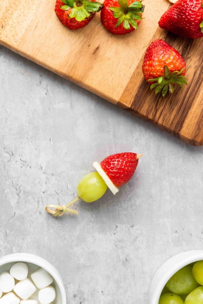 A skewer with a green grape, white cheese, and a strawberry—perfect Grinch Fruit Kabobs—rests on a gray surface. Nearby are bowls of grapes and marshmallows, with whole strawberries on a wooden cutting board.