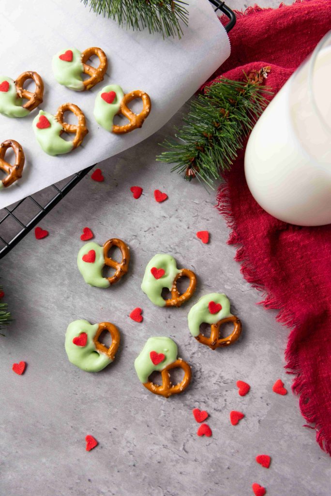Grinch Heart Pretzels—green chocolate-dipped-pretzels-for-christmas with red heart sprinkles—are arranged on a gray surface, accompanied by a glass of milk, fir branches, a red cloth, and more pretzels cooling in the background.
