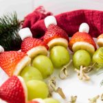 A plate of festive Grinch Fruit Kabobs made with green grapes, banana slices, strawberries, and mini marshmallows arranged to look like Santa hats, with holiday greenery and a red napkin in the background.