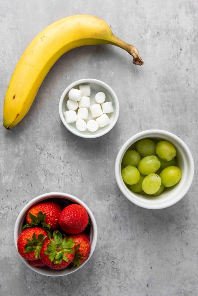 A banana, a bowl of mini marshmallows, green grapes, and strawberries are arranged on a gray surface—perfect ingredients for making festive Grinch Fruit Kabobs.