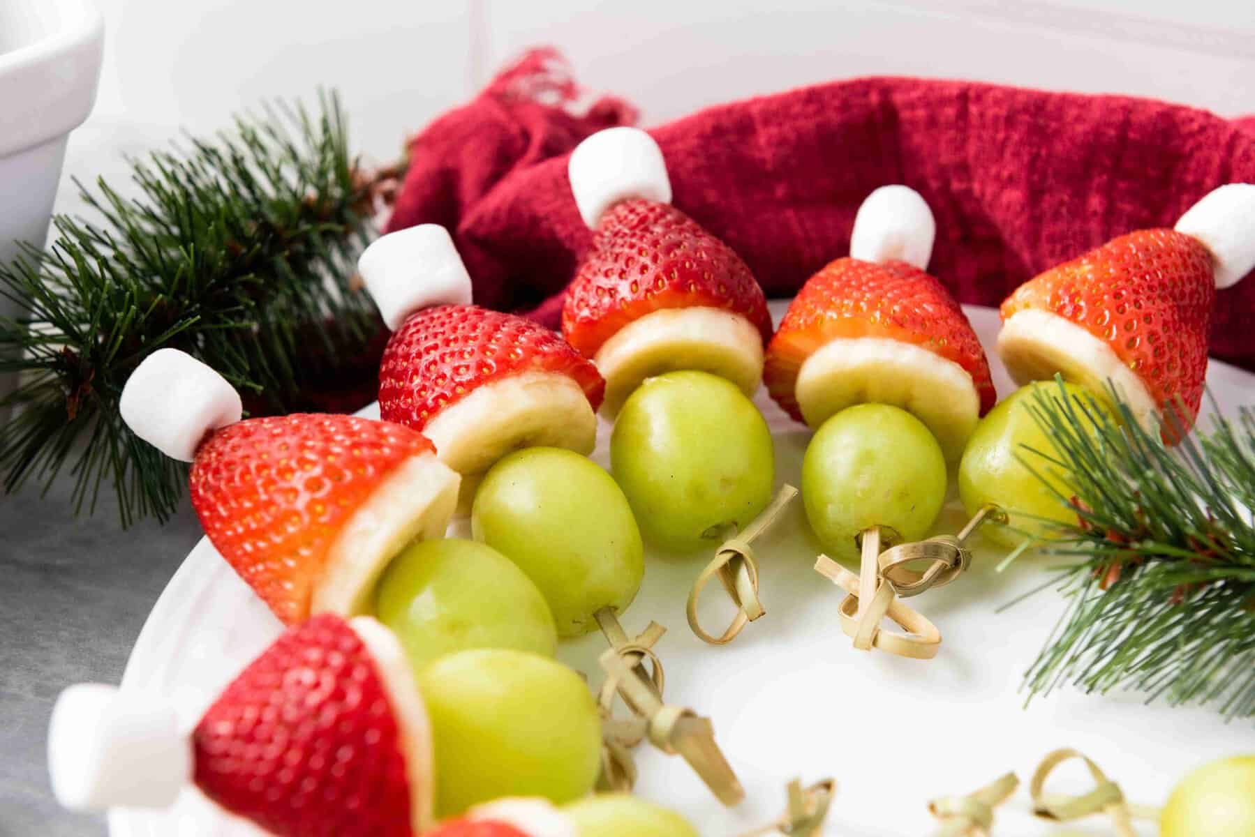 A plate of festive Grinch Fruit Kabobs made with green grapes, banana slices, strawberries, and mini marshmallows arranged to look like Santa hats, with holiday greenery and a red napkin in the background.