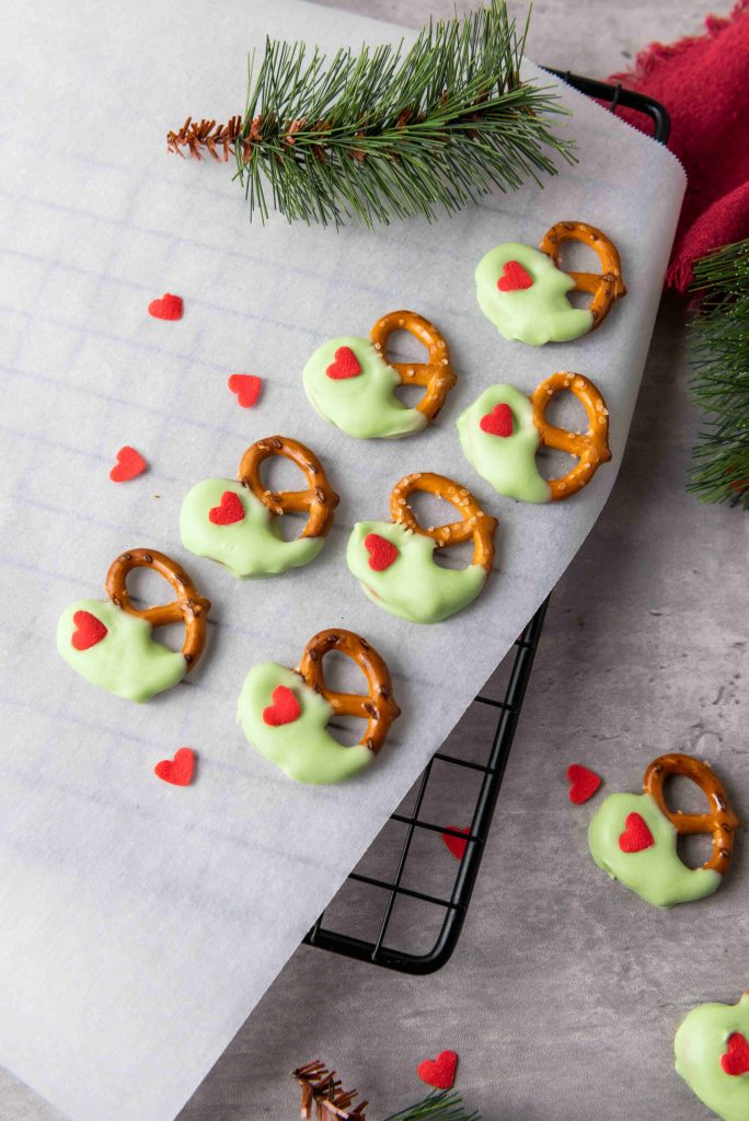 Grinch Heart Pretzels—dipped-pretzels-for-christmas—are arranged on parchment paper on a cooling rack, each coated in green chocolate and topped with a red heart, with pine branches and extra heart sprinkles nearby.