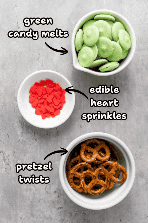 Three bowls on a gray surface display green candy melts, red heart-shaped sprinkles, and pretzel twists—perfect for making Grinch Heart Pretzels or delicious dipped-pretzels-for-Christmas. Handwritten labels and arrows identify each festive ingredient.