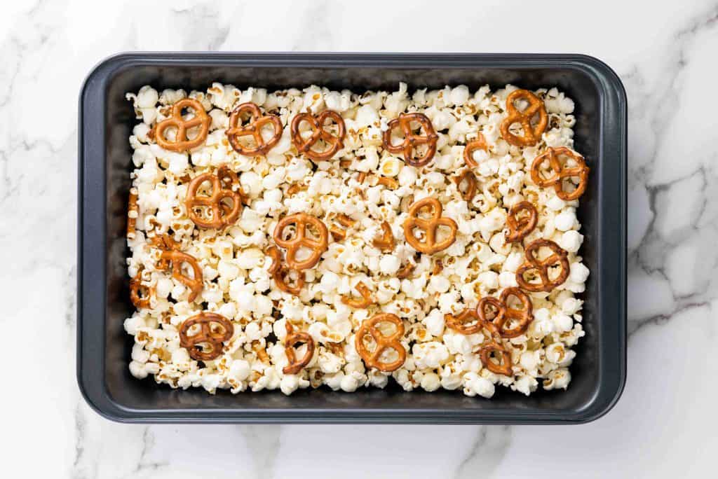 A rectangular black baking pan filled with holiday popcorn and scattered pretzels, placed on a white marble surface.