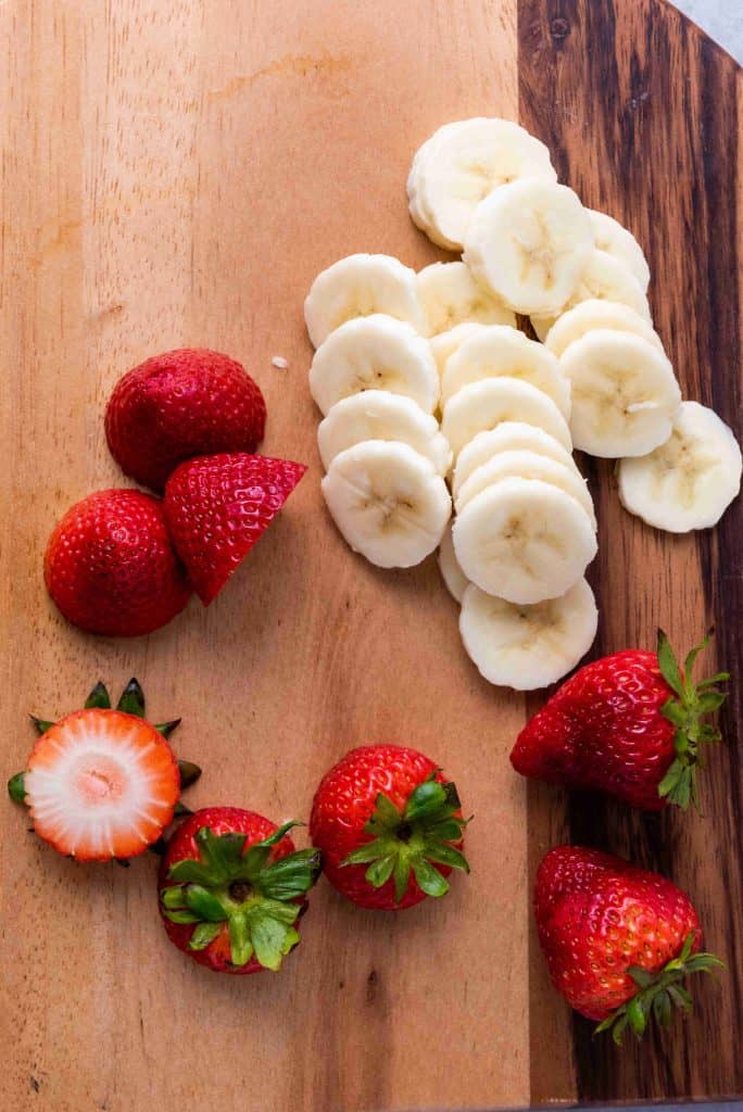 Sliced bananas and whole strawberries, some with stems, are arranged on a wooden cutting board, perfect for assembling Grinch Fruit Kabobs.