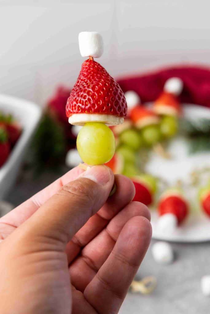 A hand holds a festive Grinch Fruit Kabob made of a green grape, banana slice, upside-down strawberry, and mini marshmallow, arranged to resemble a holiday gnome or Santa. A plate with more kabobs is in the background.