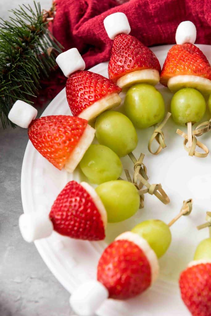 A festive plate features Grinch Fruit Kabobs—skewers of green grapes, banana slices, strawberries, and mini marshmallows arranged to resemble Christmas Santa hats. A red cloth and pine branch add seasonal charm in the background.
