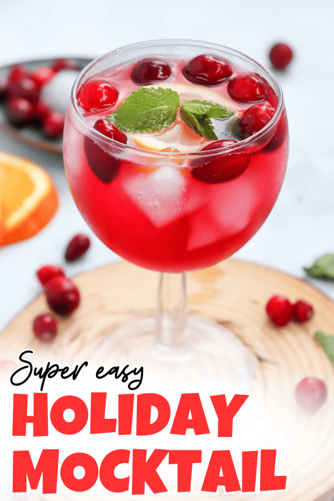A festive cranberry Christmas mocktail with ice, cranberries, and mint leaves in a stemmed glass. Text reads Super easy HOLIDAY MOCKTAIL. Orange slices and cranberries are scattered nearby.