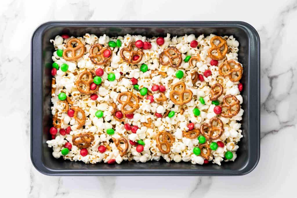 A rectangular pan filled with holiday popcorn, mini pretzels, red and green candy-coated chocolates, and white drizzle sits on a white marble surface.