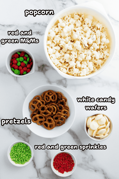 Five bowls on a marble surface contain holiday popcorn, red and green M&Ms, pretzels, white candy wafers, and festive sprinkles. Each bowl is labeled with black and white text.
