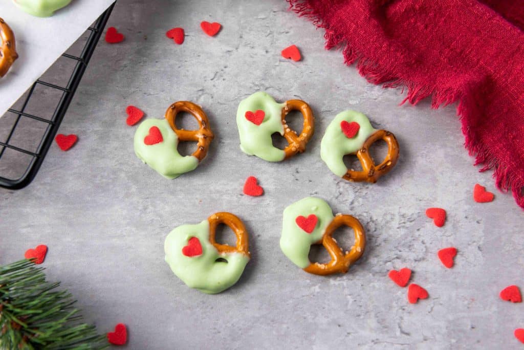 Mini Grinch Heart Pretzels, partially dipped in pale green chocolate and topped with a small red heart, are arranged on a gray surface with scattered red heart confetti and a red cloth. Perfect dipped-pretzels-for-christmas!.