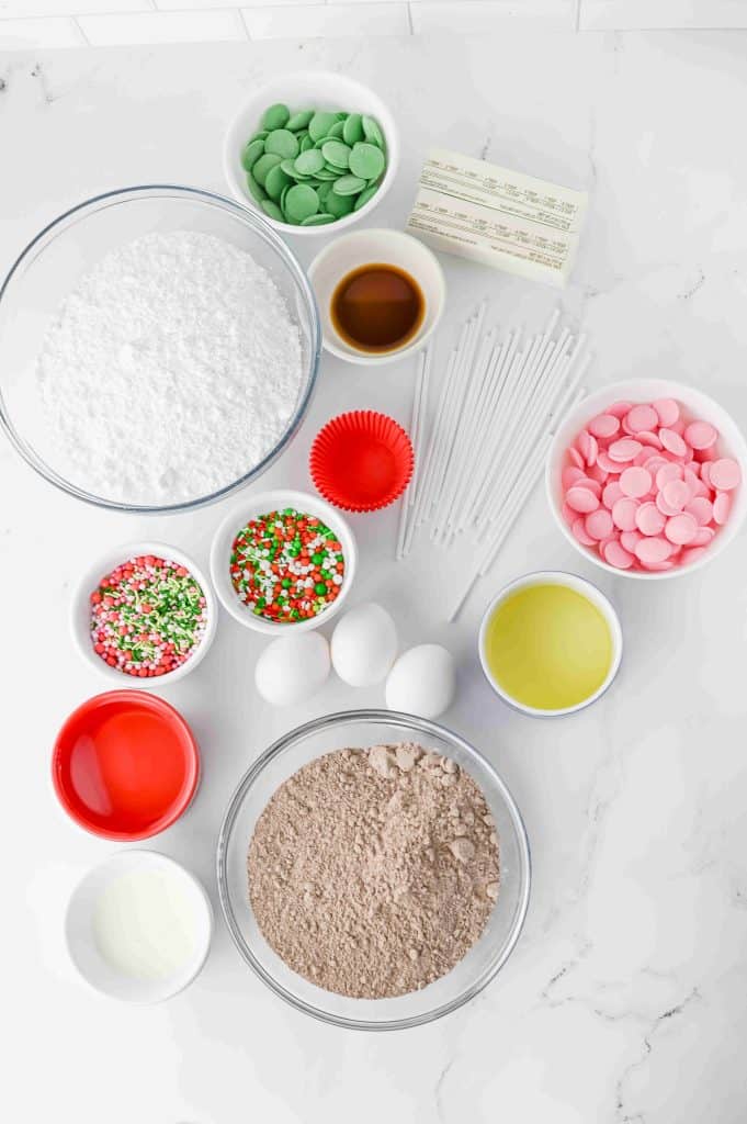 Ingredients for making cake pops are arranged on a white countertop, alongside festive elements you might use for christmas tree cupcakes, including eggs, oil, cake mix, powdered sugar, food coloring wafers, vanilla, butter, sprinkles, and cake pop sticks in separate bowls.