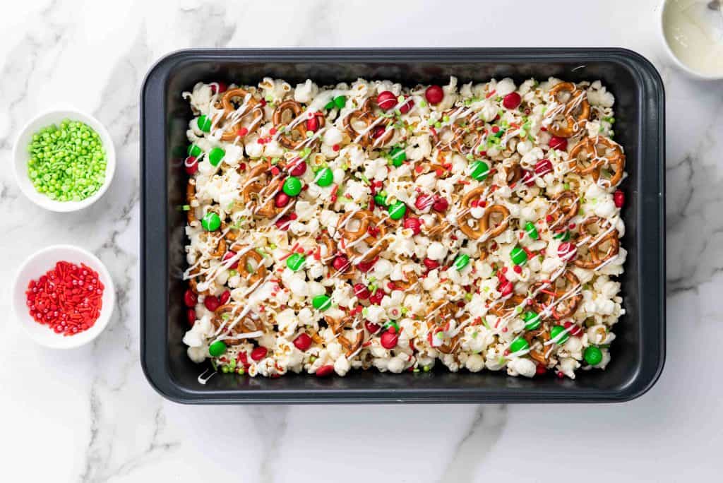 A black baking pan filled with holiday popcorn—popcorn topped with red and green candies, pretzels, festive sprinkles, and drizzled white chocolate. Two small bowls with red and green sprinkles sit nearby on a marble surface.