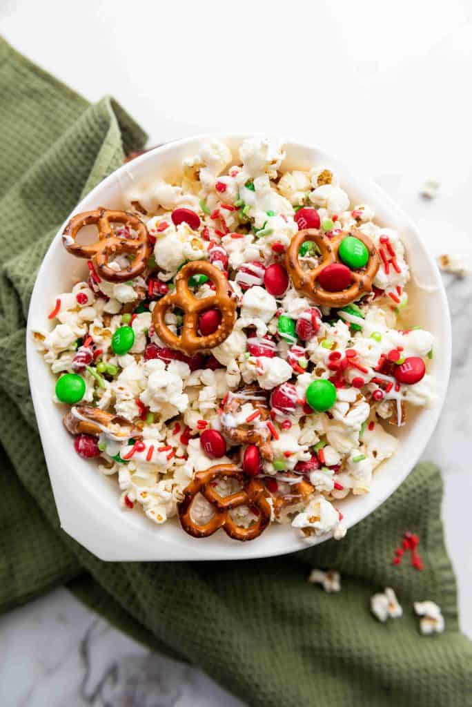 A white bowl of holiday popcorn, pretzels, red and green M&M candies, and festive sprinkles sits on a green cloth atop a white marble surface.