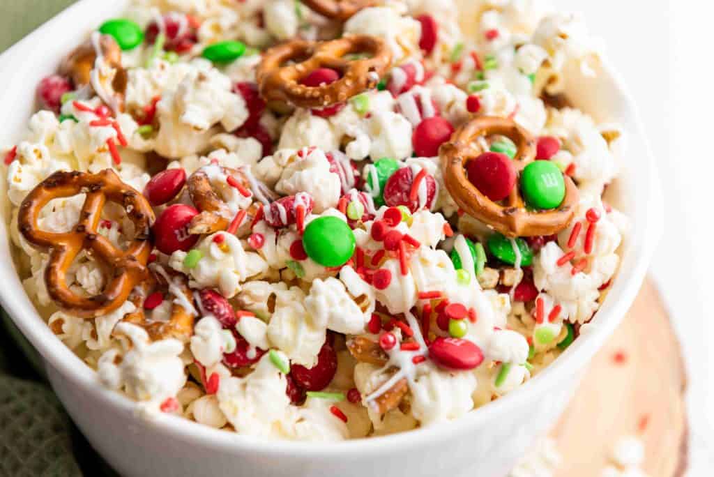 A bowl filled with holiday popcorn, red and green chocolate candies, mini pretzels, and festive sprinkles, all drizzled with white chocolate for a cheerful holiday snack mix.