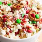 A bowl filled with holiday popcorn, red and green chocolate candies, mini pretzels, and festive sprinkles, all drizzled with white chocolate for a cheerful holiday snack mix.