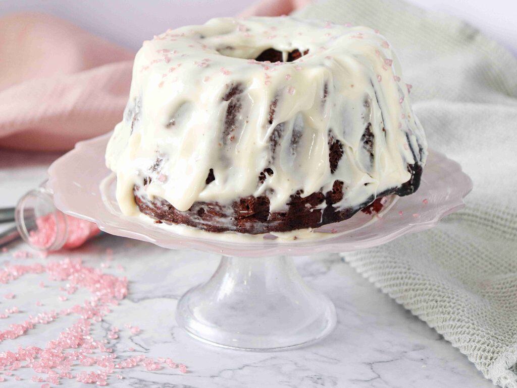 A Christmas Red Velvet Bundt Cake with white icing drips sits on a glass cake stand. Pink sugar sprinkles are scattered on the cake and table, with a pink napkin and green cloth in the background.