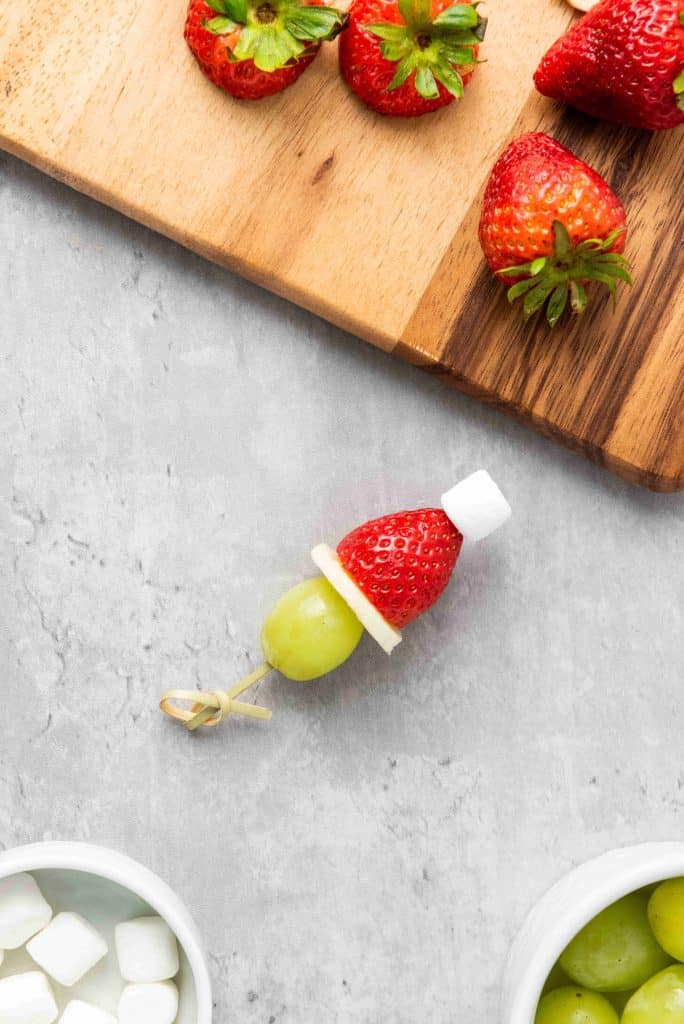 Grinch Fruit Kabobs feature a skewer with a green grape, banana slice, strawberry, and mini marshmallow for a festive holiday snack, with fresh strawberries on a wooden cutting board nearby.