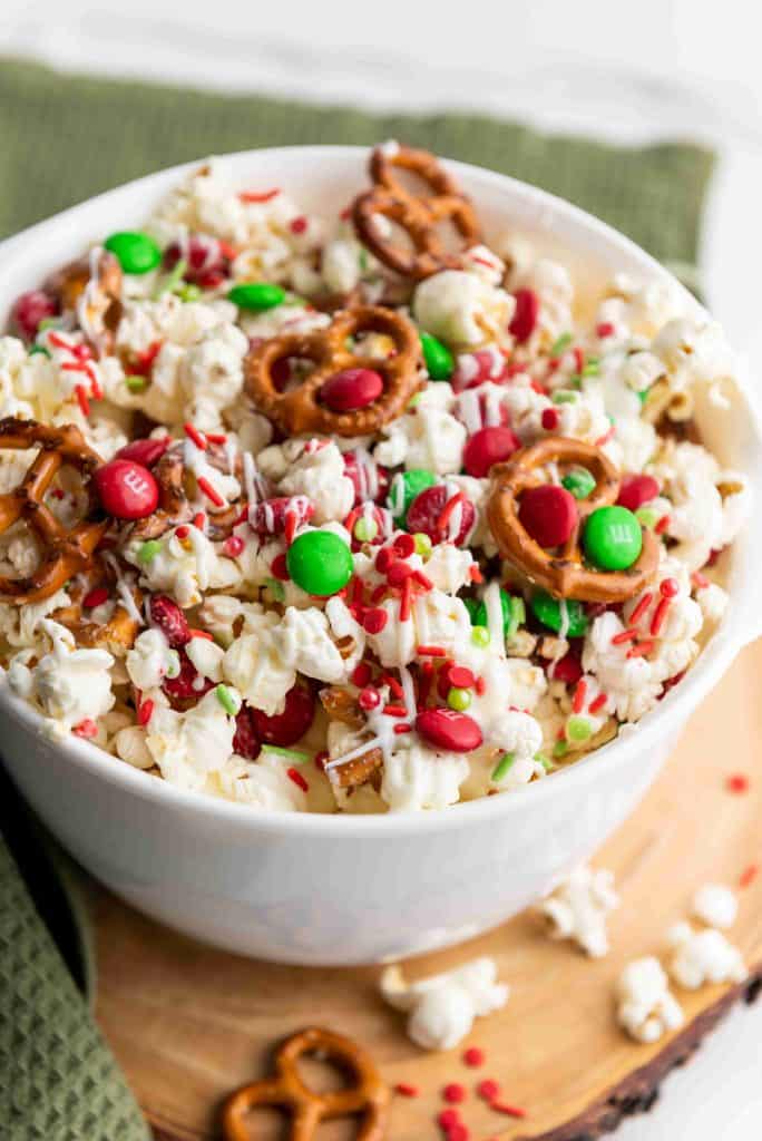A white bowl filled with holiday popcorn—popcorn, mini pretzels, red and green candy-coated chocolates, and festive sprinkles—all drizzled with white chocolate, sits on a wooden surface with a green napkin.