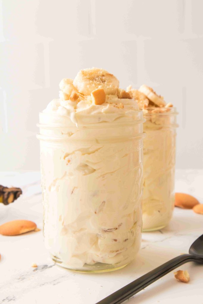 Two glass jars filled with creamy Christmas Banana Pudding, topped with banana slices and crumbled cookies, sit on a white surface with vanilla wafers scattered nearby. A black spoon rests in the foreground.