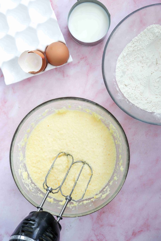 A mixing bowl with batter and an electric hand mixer, surrounded by a bowl of flour, a cup of milk, eggshells, and whole eggs on a pink surface—perfect for preparing a Valentine's Day cake.