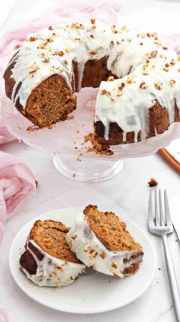 A Christmas Carrot Cake with white icing and chopped nuts sits on a glass stand. Two slices of the cake are served on a small white plate next to a fork, with pink fabric and crumbs scattered around.
