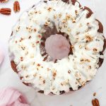 A round Christmas Carrot Cake bundt with white icing and chopped pecans on top, surrounded by cinnamon sticks, pecans, and a light pink cloth on a white surface.