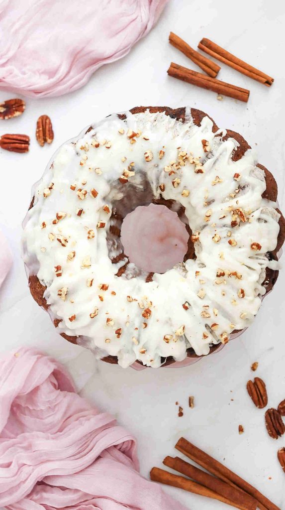 A round Christmas Carrot Cake bundt with white icing and chopped pecans on top, surrounded by cinnamon sticks, pecans, and a light pink cloth on a white surface.