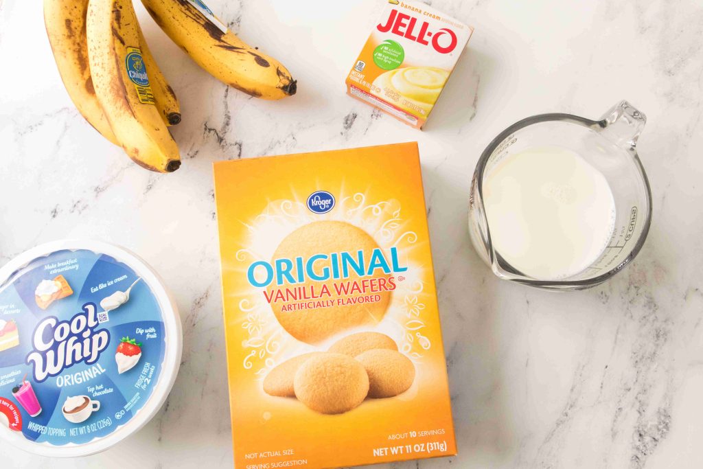 Top-down view of ingredients for Christmas Banana Pudding on a white surface: bananas, Cool Whip container, vanilla wafers box, vanilla pudding mix packet, and a measuring cup filled with milk.