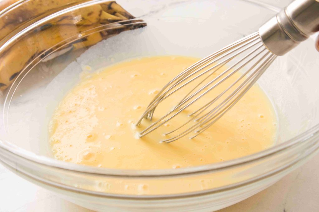 A metal whisk mixing a light yellow Christmas Banana Pudding batter in a clear glass bowl, with ripe bananas in the background on a white surface.