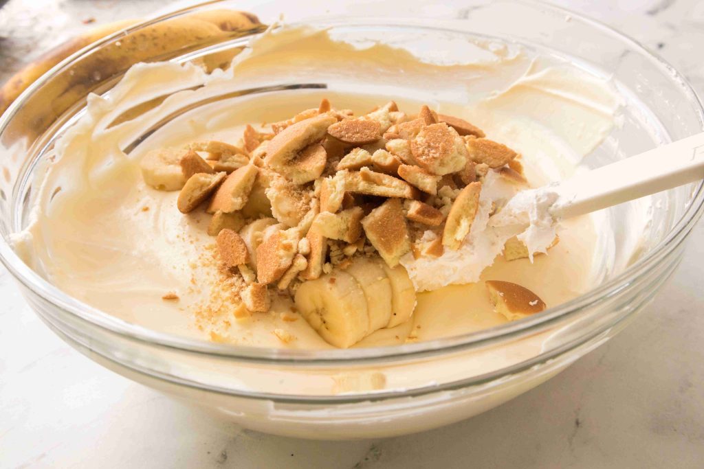 A glass bowl filled with creamy Christmas Banana Pudding, layered with sliced bananas and crushed vanilla wafers, with a spatula resting on the side.