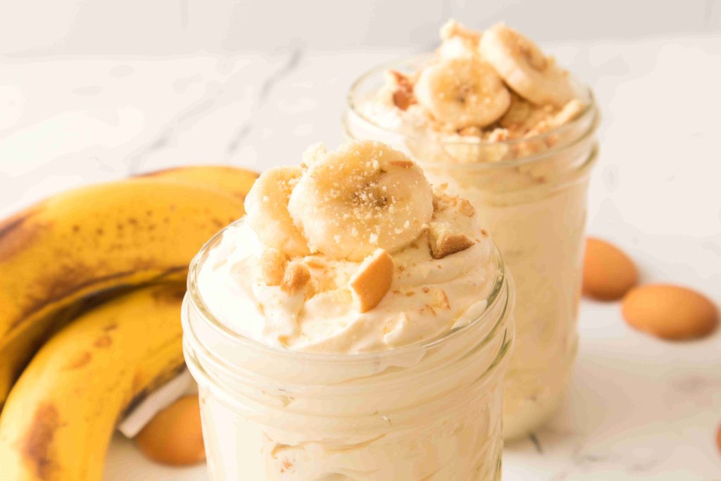 Two glass jars filled with creamy Christmas Banana Pudding, topped with banana slices and crumbled cookies. In the background, ripe bananas and vanilla wafer cookies are visible on a light surface.