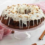 A Christmas Carrot Cake with white icing and chopped nuts sits on a glass cake stand, surrounded by cinnamon sticks, pecans, and pink fabric on a light surface.