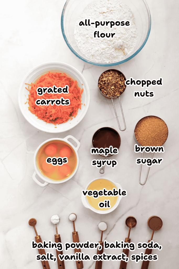 Top-down view of ingredients for Christmas Carrot Cake, featuring bowls of flour, grated carrots, chopped nuts, eggs, maple syrup, brown sugar, vegetable oil, and measuring spoons with powders and festive spices.