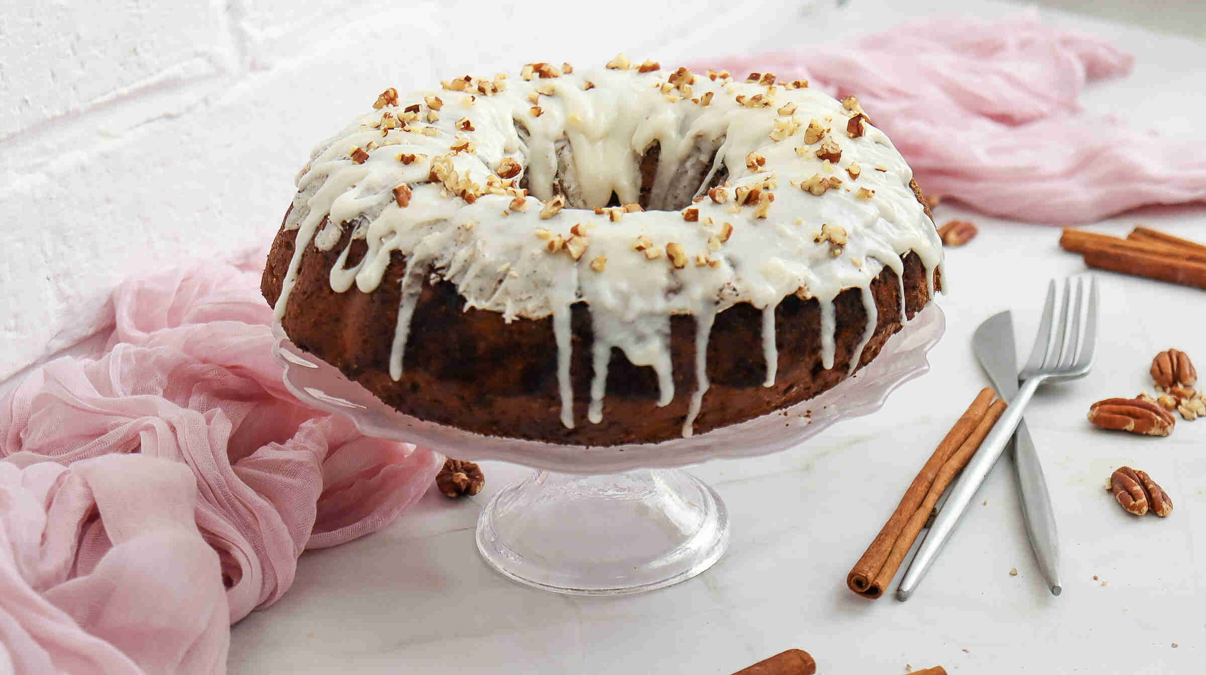 A Christmas Carrot Cake with white icing and chopped nuts sits on a glass cake stand, surrounded by cinnamon sticks, pecans, and pink fabric on a light surface.